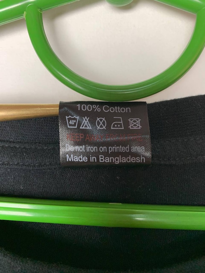 Puede incluir: Primer plano de una etiqueta de ropa negra con texto blanco. La etiqueta dice "100% Algod&oacute;n" e incluye s&iacute;mbolos de cuidado para lavar y planchar. Tambi&eacute;n dice "No planchar sobre la zona estampada" y "Hecho en Bangladesh."
