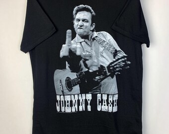 May include: Black t-shirt featuring a black and white image of Johnny Cash giving the middle finger while playing a guitar. The text "JOHNNY CASH" is printed in white below the image. The shirt has a small gold tag with the text "FINN MC LONDON®" on the top.
