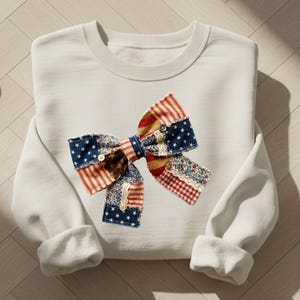 Peut inclure: Un sweat-shirt gris clair orné d'un grand nœud décoratif. Le nœud est composé de différents patchworks de tissus aux motifs rouges, blancs et bleus, avec des étoiles et des rayures. Le nœud est agrémenté de petits boutons.