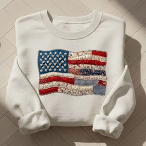 May include: White sweatshirt featuring a patchwork design of the American flag. The flag design incorporates blue, red, and white fabrics with floral and gingham patterns. The sweatshirt has a crew neck and long sleeves.