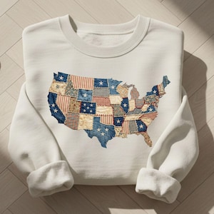 May include: A white sweatshirt featuring a patchwork design of the United States map. The map is composed of various fabric patterns in red, white, and blue, with star accents. The sweatshirt has a crew neck and long sleeves.