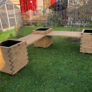 May include: A wooden planter bench with two square planters linked by a slatted walkway. The planters and bench are made of light-coloured wood and sit on a grassy lawn. The planters have black interiors.