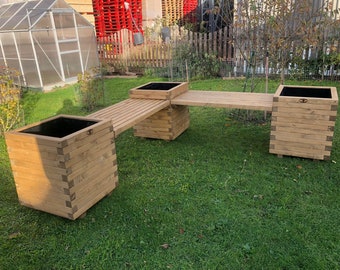 May include: A wooden planter bench with two square planters linked by a slatted walkway. The planters and bench are made of light-coloured wood and sit on a grassy lawn. The planters have black interiors.
