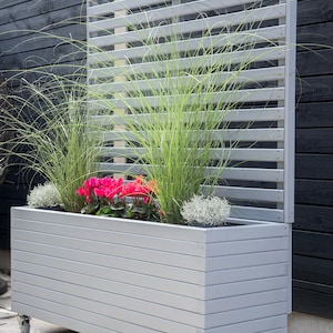 May include: A grey wooden planter with a slatted screen, filled with green grasses, pink flowers, and silver plants. The planter has wheels. The planter is designed for outdoor use and is approximately 120cm long.