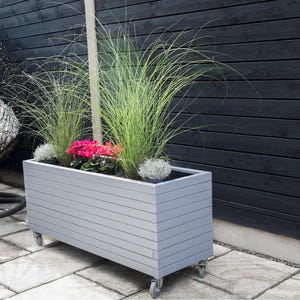 May include: A grey wooden planter box with a slatted design sits on a patio. The planter is filled with green grass and pink flowers. The planter has wheels on the bottom.