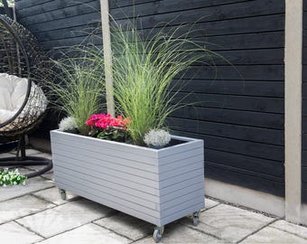 May include: A grey wooden planter box with a slatted design sits on a patio. The planter is filled with green grass and pink flowers. The planter has wheels on the bottom.