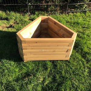 May include: A triangular wooden planter box with a natural wood finish. The planter is empty and sits on a grassy surface.