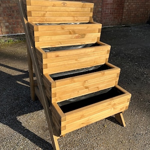May include: A wooden vertical garden planter with five tiers. The planter is made of natural wood and has a dark brown stain. The planter is standing on a gravel surface.