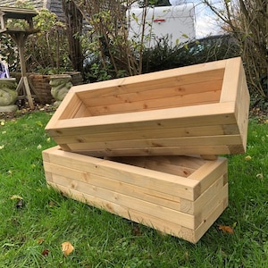 May include: Two unfinished wooden planter boxes stacked on top of each other. The boxes are made of natural wood and have a rectangular shape.