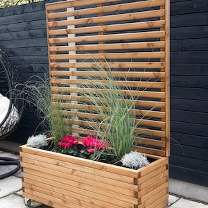 May include: A wooden planter box with a slatted privacy screen. The planter is filled with pink flowers, tall green grass, and small silver plants. The planter box is on wheels and is made of light brown wood.
