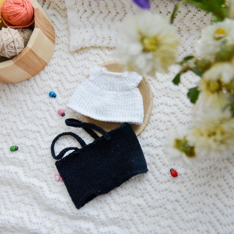 May include: Two knitted doll clothes, one white and one black, on a white crocheted surface. The white dress has short sleeves and a round neckline. The black dress has thin straps and a straight neckline.