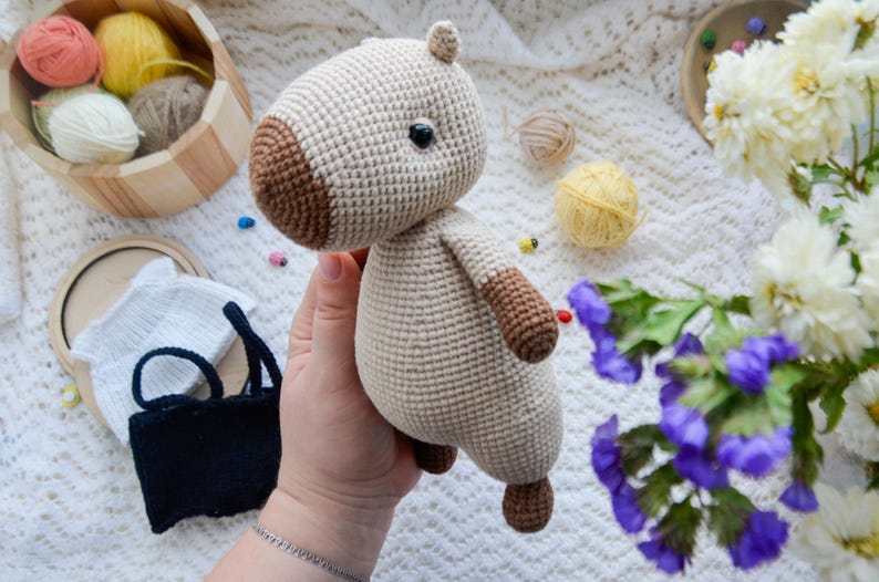 May include: A crocheted stuffed animal in the shape of a horse. The horse is beige with brown accents and has a black eye. The horse is being held in a person's hand.