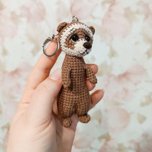 May include: A handmade crochet keychain featuring a brown and white ferret. The ferret has a detailed design with a white stripe across its head and a silver keychain ring attached. The ferret is standing upright.