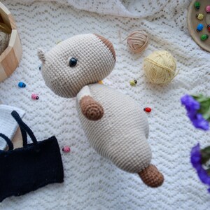 May include: A crocheted stuffed animal in the shape of a dog. The dog is beige with brown accents and has a white crocheted shirt on. The dog is lying on a white crocheted blanket.