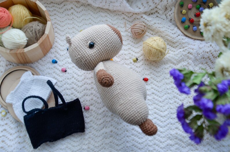 May include: A crocheted stuffed animal in the shape of a dog. The dog is beige with brown accents and has a white crocheted shirt on. The dog is lying on a white crocheted blanket.