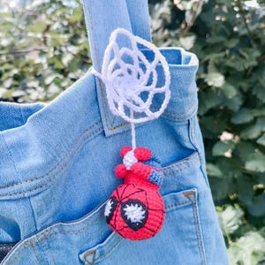 May include: A light blue denim tote bag featuring a small, handmade crochet charm. The charm consists of a white crocheted spider web attached to a red and blue crocheted character head with large white eyes, hanging from the bag's shoulder strap.