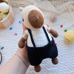 May include: A crocheted stuffed animal with brown and white fur, wearing a white shirt and black overalls. The animal is being held by a hand.