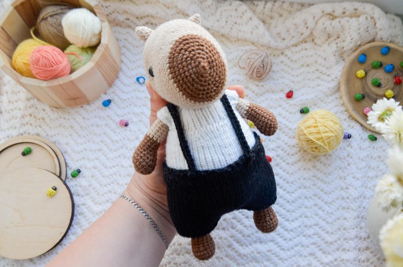 May include: A crocheted stuffed animal with brown and white fur, wearing a white shirt and black overalls. The animal is being held by a hand.