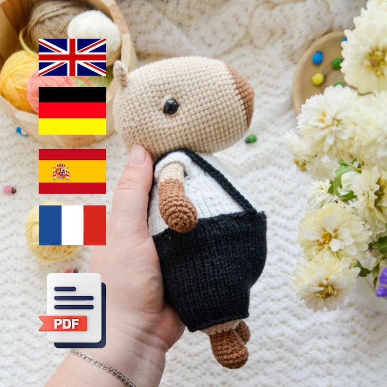 May include: Crocheted amigurumi toy of a brown and white capybara wearing a white shirt and black overalls. The toy is being held in a hand. The image also includes flags of the United Kingdom, Germany, Spain, and France, and a PDF icon.