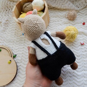 May include: A crocheted stuffed animal in the shape of a bear wearing a white shirt with black suspenders and black pants. The bear is brown and beige and has a button on its shirt.
