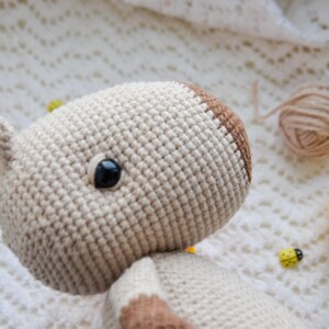 May include: Close-up of a crocheted stuffed animal with brown and white yarn. The animal has one black eye and is lying on a white crocheted blanket.