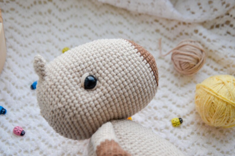 May include: Close-up of a crocheted stuffed animal with brown and white yarn. The animal has one black eye and is lying on a white crocheted blanket.