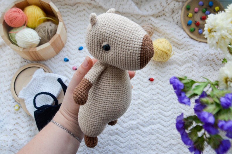 May include: A crocheted stuffed animal in the shape of a capybara. The capybara is beige with brown accents and has a black eye.