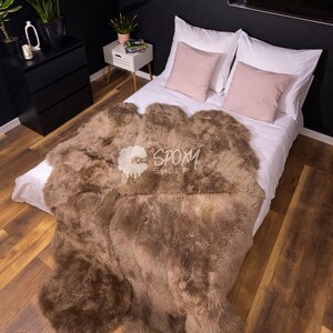 May include: A brown sheepskin rug draped over a bed with white linens and pink pillows. The rug has a thick, textured appearance and covers a significant portion of the bed. The room has dark walls and wooden flooring.