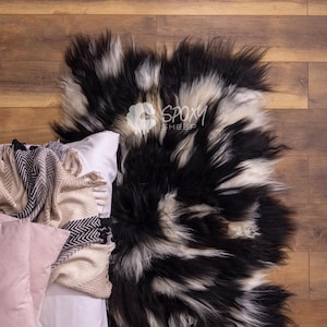Könnte beinhalten: Ein schwarz-weißer Schaffellteppich mit langem, zotteligem Fell. Der Teppich liegt auf einem Holzboden neben einem Bett mit Kissen und Decken. Der Teppich hat eine natürliche, organische Form und Textur.