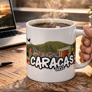 Può includere: Tazza da caffè bianca con un design di Caracas, piena di caffè nero. La tazza presenta un'illustrazione colorata dello skyline della città e la parola "Caracas" in un font stilizzato. Una mano tiene la tazza, con un laptop e una penna sullo sfondo.