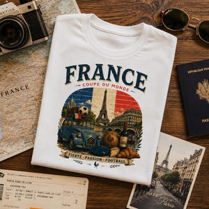 May include: A white t-shirt featuring a graphic print with the text FRANCE COUPE DU MONDE and FIERT&Eacute; PASSION FOOTBALL. The design includes a blue car, a soccer ball, the Eiffel Tower, a bottle of wine, bread, and a croissant against a French flag.