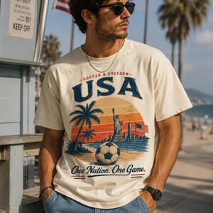 May include: Cream-colored t-shirt with a graphic featuring a soccer ball, palm trees, the Statue of Liberty, and the text "USA Soccer & Freedom." The shirt also includes the phrase "One Nation. One Game."