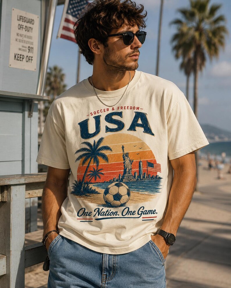 May include: Cream-colored t-shirt with a graphic featuring a soccer ball, palm trees, the Statue of Liberty, and the text "USA Soccer & Freedom." The shirt also includes the phrase "One Nation. One Game."