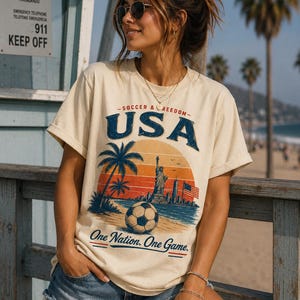 May include: Cream-colored t-shirt with a graphic featuring "USA" in blue, a soccer ball, palm trees, and the Statue of Liberty. The text "One Nation. One Game." is below the graphic. The shirt is worn with denim shorts.