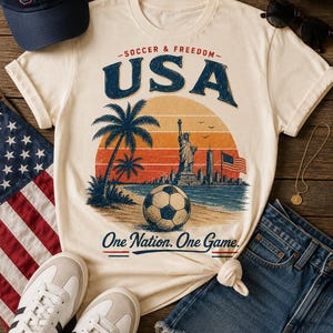 May include: Cream-colored t-shirt with a graphic of a soccer ball on a beach with palm trees, the Statue of Liberty, and the text "USA". A navy blue baseball cap and denim shorts are also visible.