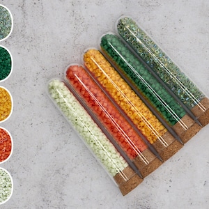 Könnte beinhalten: Sechs Glasröhrchen gefüllt mit bunten Perlen. Die Röhrchen sind in einer Reihe angeordnet, wobei das Korkende jedes Röhrchens zum Betrachter zeigt. Die Perlen sind in den folgenden Farben: grün, gelb, orange, rot und weiß.