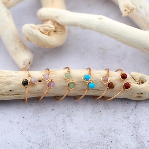 Könnte beinhalten: Ein Satz von sieben goldenen, drahtgewickelten Ringen mit verschiedenen Edelsteinen. Die Ringe sind auf einer hellbraunen Holzoberfläche angeordnet.