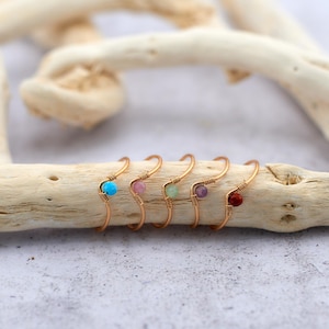 Könnte beinhalten: Fünf goldene Drahtringe mit unterschiedlich farbigen Edelsteinen. Die Ringe sind in einer Reihe auf einem Stück Treibholz angeordnet.