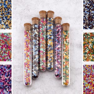 Könnte beinhalten: Sechs Glasröhrchen gefüllt mit bunten Perlen. Die Perlen sind in verschiedenen Farben, darunter Rot, Orange, Gelb, Grün, Blau und Lila. Die Röhrchen sind in einer Reihe angeordnet, wobei die Korkstopfen zum Betrachter zeigen.