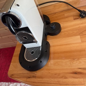 Könnte beinhalten: Eine weiß-silberne Espressomaschine mit schwarzem Sockel und transparentem Wassertank. Die Maschine steht auf einer Holzoberfläche, mit einem schwarzen Netzkabel und Stecker. Sie hat ein schlankes, modernes Design.