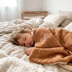 Peut inclure: Un enfant endormi est couvert d'une couverture marron à pois blancs et le nom "Mance" brodé en blanc. La couverture a une doublure en sherpa blanche et douce. L'enfant est sur un lit avec une literie aux tons neutres.
