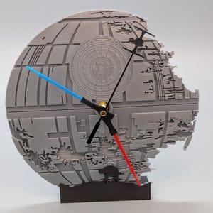 May include: A gray Death Star clock with a black base. The clock hands are designed as a blue lightsaber and a red lightsaber. The clock face has detailed surface features.
