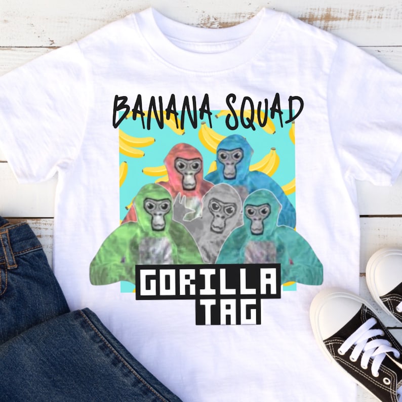 K&ouml;nnte beinhalten: Wei&szlig;es T-Shirt mit einem Grafikdruck von f&uuml;nf Cartoon-Gorillas, die verschiedene farbige Shirts tragen und von gelben Bananen umgeben sind. Der Text "Banana Squad" befindet sich &uuml;ber den Gorillas und "Gorilla Tag" darunter.