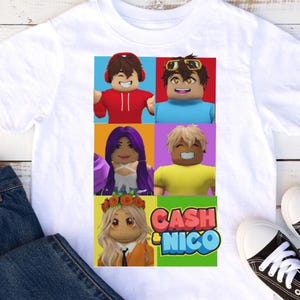 May include: White t-shirt featuring a colorful graphic of six cartoon characters in square panels. The graphic includes the text "CASH & NICO" in a green and blue design. The shirt is displayed on a white surface with blue jeans and black and white sneakers.