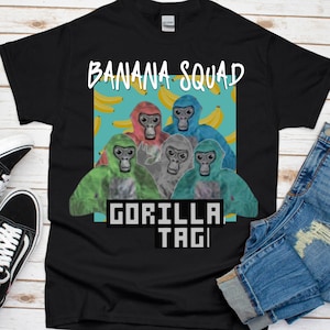 K&ouml;nnte beinhalten: Schwarzes T-Shirt mit einem Grafikdruck von vier bunten Gorillas und dem Text "Banana Squad Gorilla Tag". Die Gorillas sind von gelben Bananen auf blauem Hintergrund umgeben.