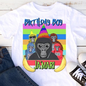May include: White t-shirt with a colorful graphic of monkeys wearing party hats and sunglasses. The text "Birthday Boy" is above the monkeys and the text "Ja'Mari" is below the monkeys. Two yellow bananas are on either side of the text.