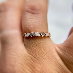 May include: A gold ring with a repeating pattern of white, oval-shaped stones and small, clear gemstones. The ring is set on a finger, showcasing its delicate design and intricate details. The ring's band is a warm gold color.