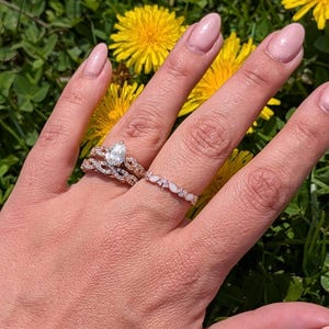 May include: A hand wearing three rings: a pear-shaped diamond engagement ring, a twisted band, and a delicate band with opal-like stones. The rings are set against a backdrop of green grass and yellow flowers.