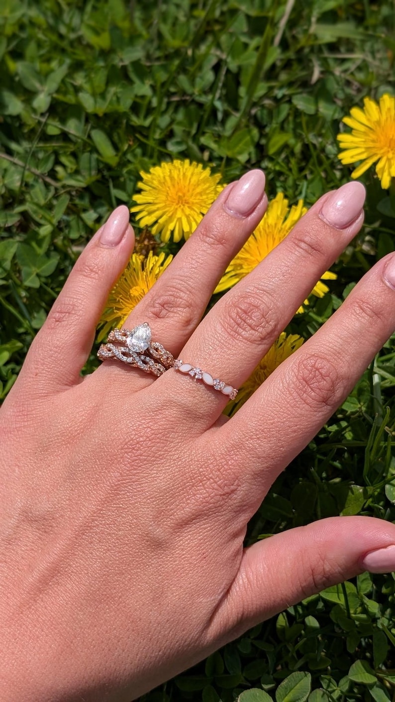 May include: A hand wearing three rings: a pear-shaped diamond engagement ring, a twisted band, and a delicate band with opal-like stones. The rings are set against a backdrop of green grass and yellow flowers.