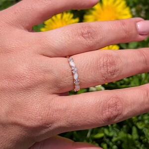 May include: A gold-toned ring with alternating oval-shaped white gemstones and small clear stones. The ring is worn on a hand with pink nail polish, set against a backdrop of green grass and yellow flowers.